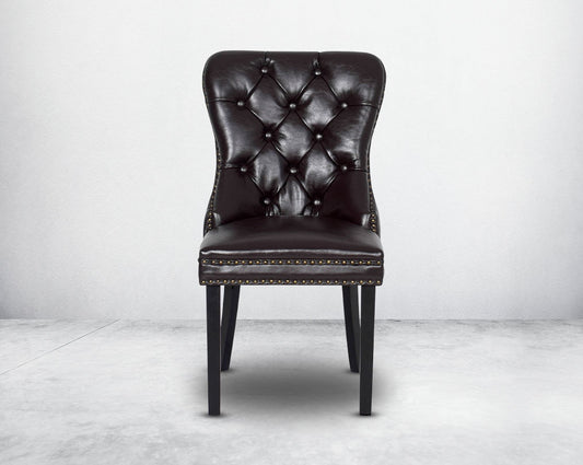 Dining chair upholstered in faux leather with front view buttoned tufted back the chair has antique gold rivets, legs in dark vintage brown wood.