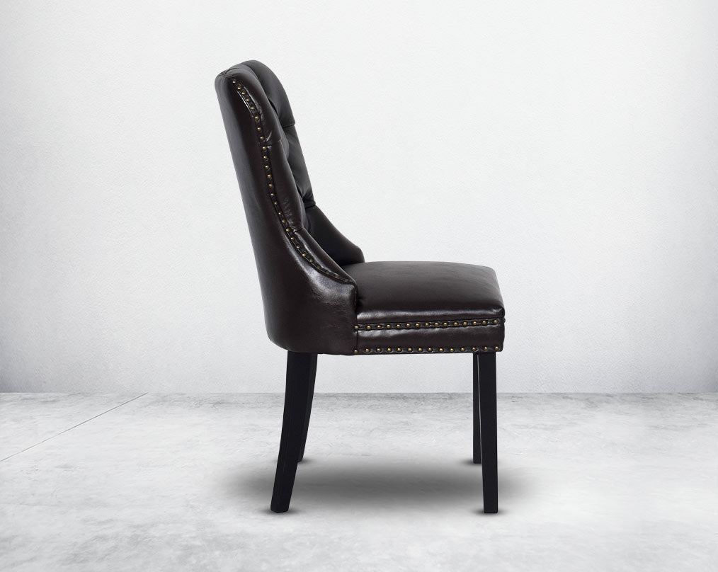 Dining chair upholstered in faux leather with side view buttoned tufted back the chair has antique gold rivets, legs in dark vintage brown wood.