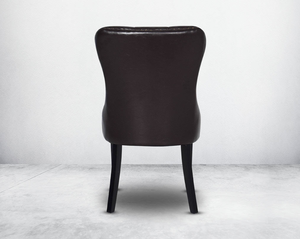 Dining chair upholstered in faux leather with back view buttoned tufted back the chair has antique gold rivets, legs in dark vintage brown wood.