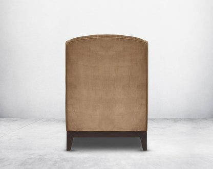 Wing arm chair upholstered in fabric and tufted with antique gold rivets on the arms and front with cushion and legs in brown oak back view