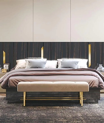 A king-sized bed with a dark walnut veneer headboard with hydraulic lift storage and gold-finished stainless-steel inlay trim. The bed is accompanied by a high gloss lacquered bedside table with a drawer and a dresser with multiple drawers, both with gold-finished stainless steel trim and wood bases.